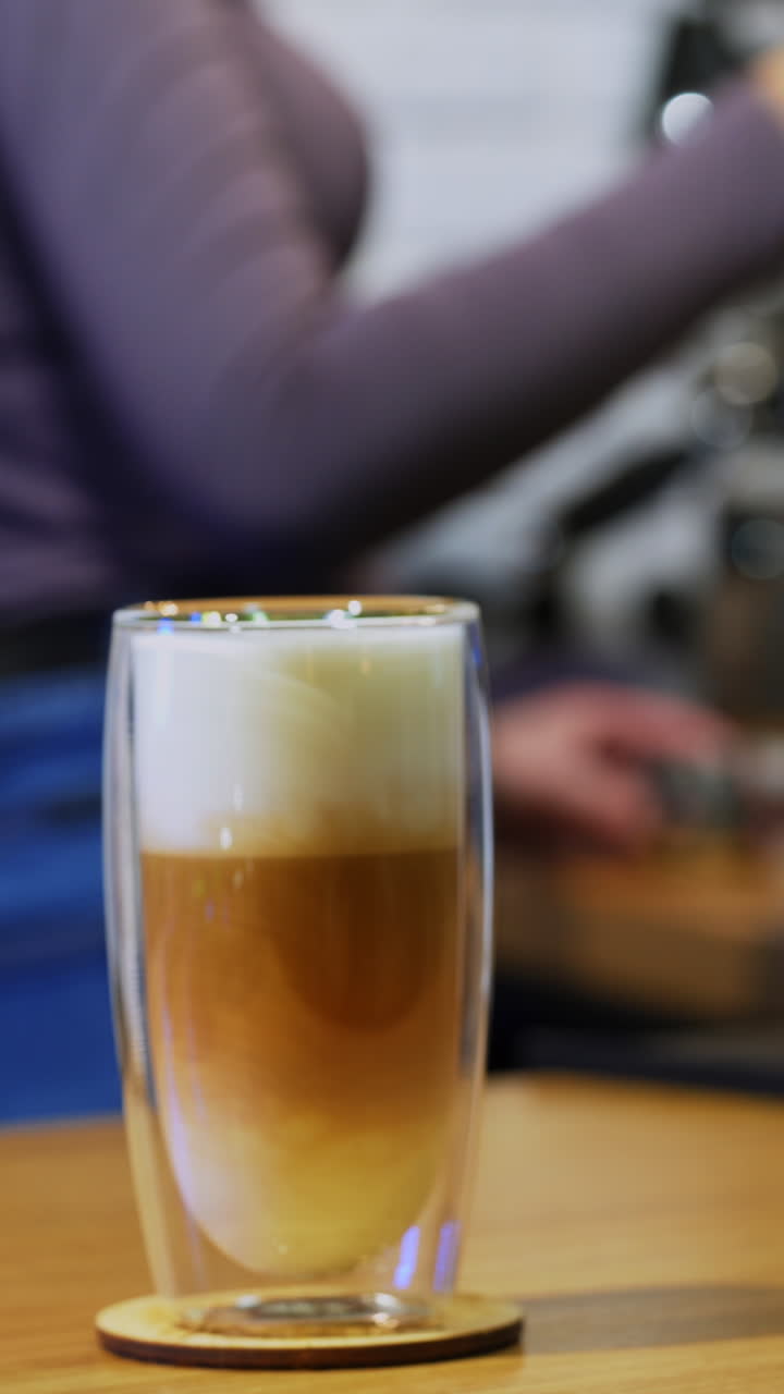 Cappuccino or latte in glass. Woman puts drink from coffee machine. Fresh coffee with whipped cream in tall clear glass on table. Close-up. Vertical video