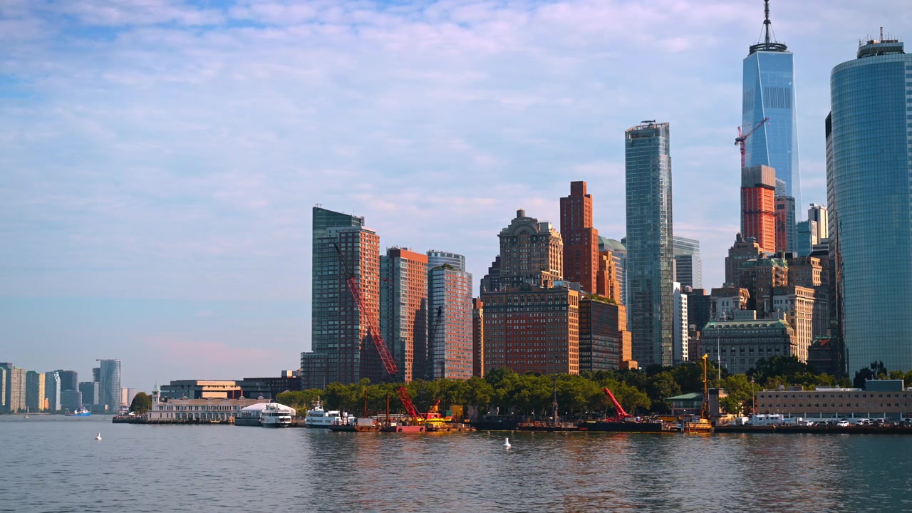 Horizonte de Lower Manhattan acima da orla do Rio Hudson. Guindastes e torres variadas captam a luz solar quente sob um céu parcialmente nublado.