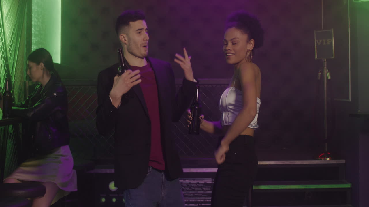 feliz pareja multiétnica bebiendo cerveza y bailando en la discoteca, mientras una chica seria sentada en un taburete y usando teléfono móvil