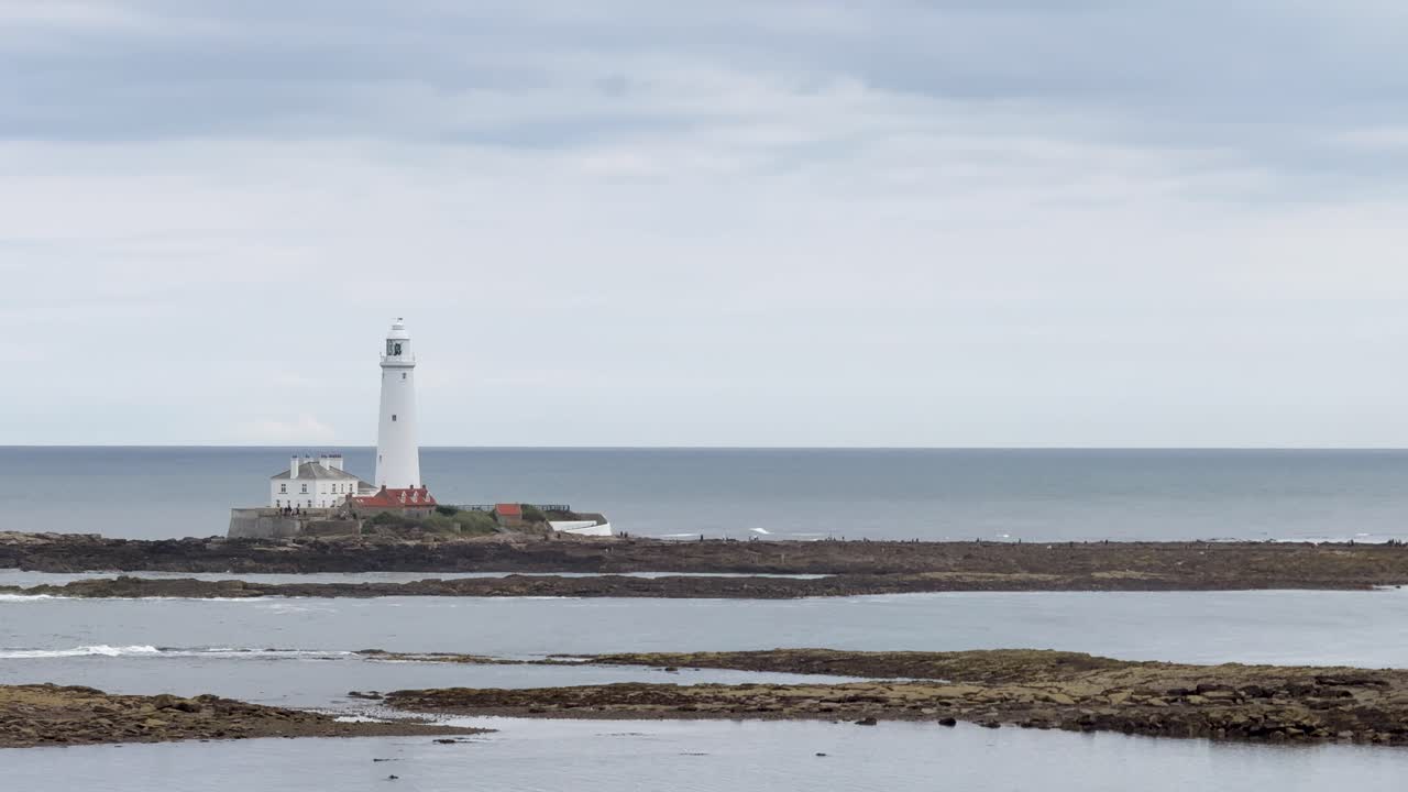 A stationary camera captures a distant lighthouse on a rocky island under cloudy skies, with calm sea and muted daylight, emphasizing tranquil coastal scenery