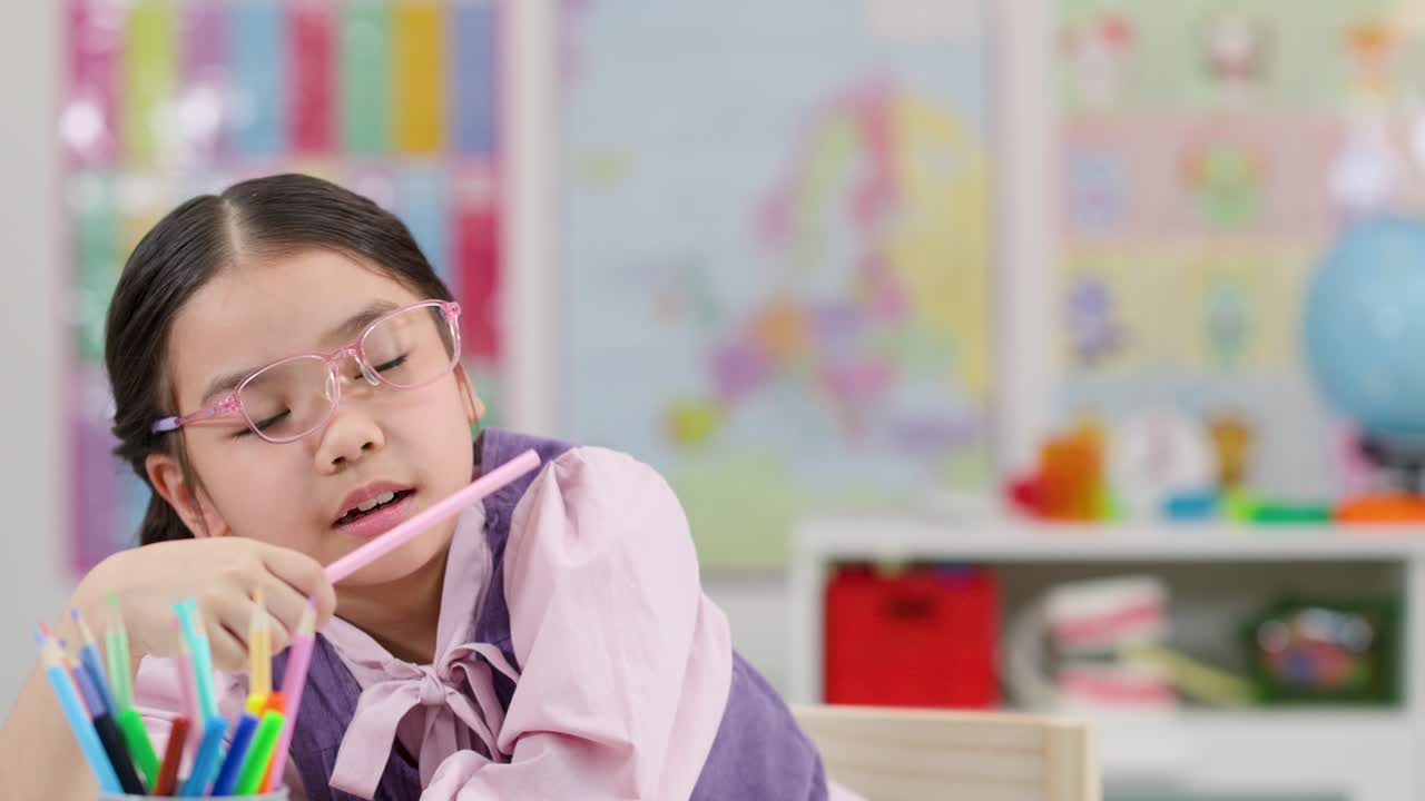 Una studentessa asiatica sorridente e allegra aggiusta gli occhiali seduta a un banco colorato in classe, circondata da matite