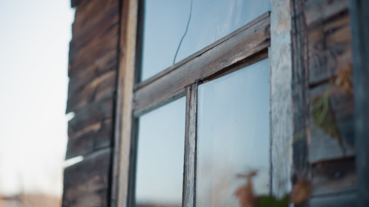 Værslitt trevindu på husets eksteriør, nærbilde av flassende maling, teksturert ramme og reflekterende glass i skumringen, uskarpt løvverk i forgrunnen fremkaller landlig nostalgi og en rolig atmosfære