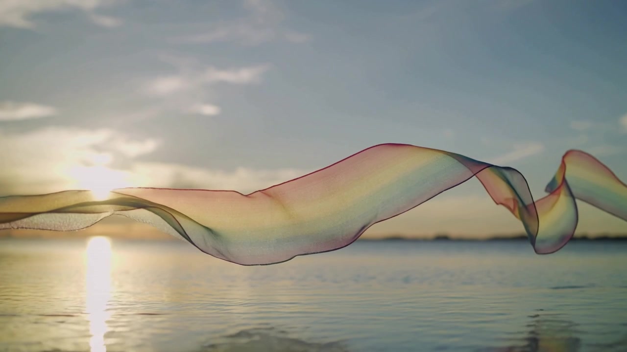 Gliding rainbow ribbon riding gentle breeze into arched crest over calm water at sunrise reflection