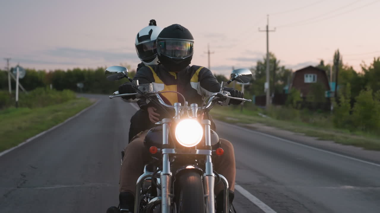 Evening motorcycle rider with passenger cruising on rural road, bright headlight glowing as they approach bend, road sign visible at side, highlighting traffic, safety and freedom