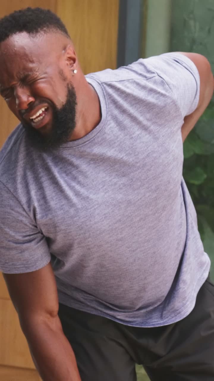 Vertical video: Doubling over from pain, man in grey tee rubbing lower back on porch by glass door