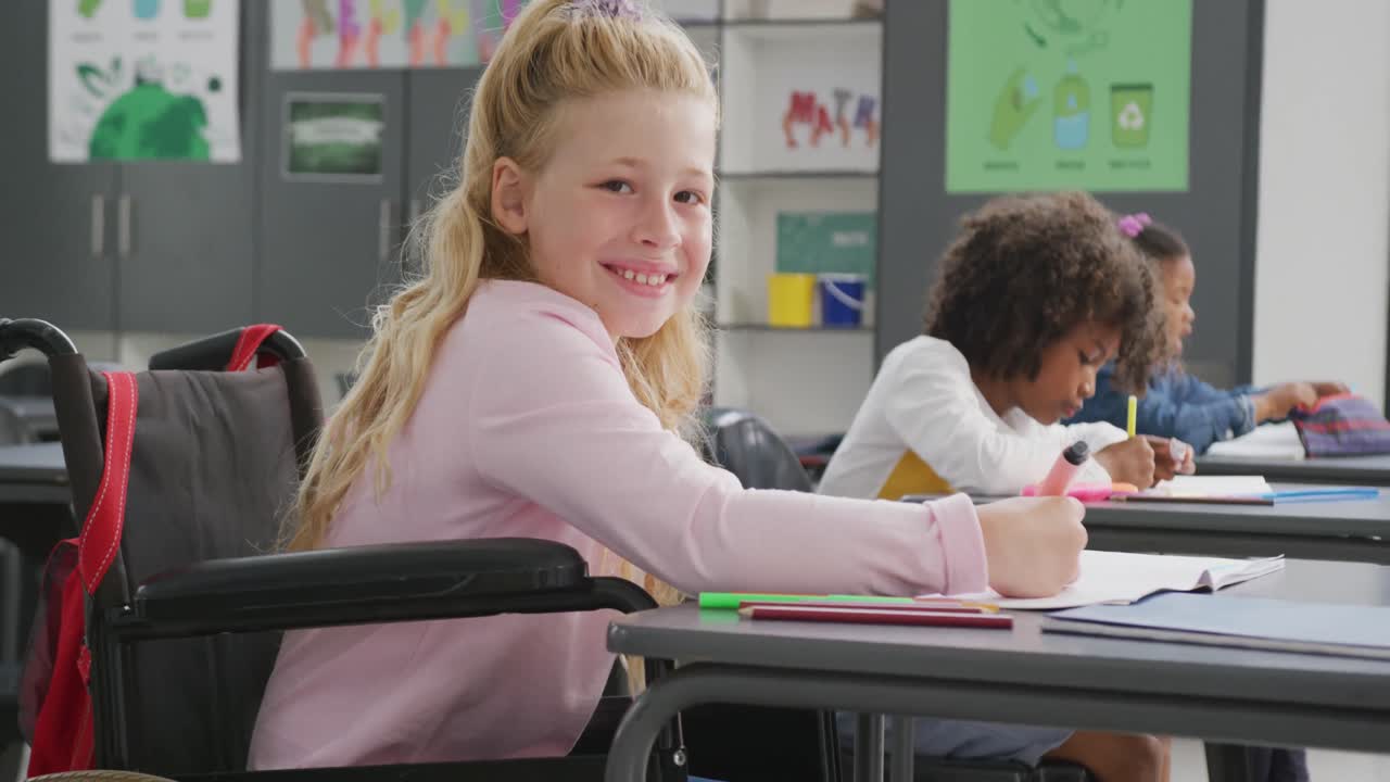 Video portrait of smiling caucasian schoolgirl in wheelchair at desk in diverse class