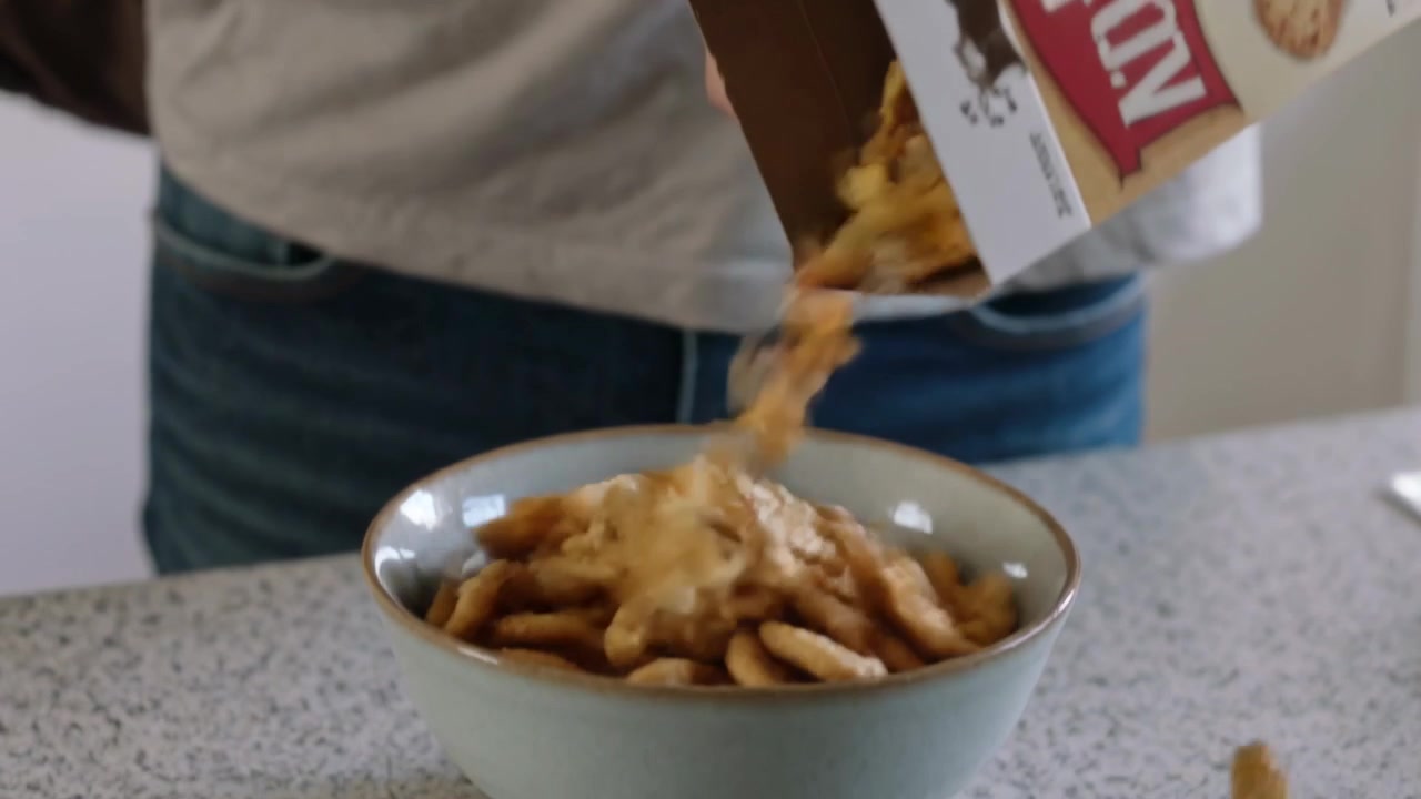 Homem chegando com mochila, despejando cereal da caixa na tigela na bancada da cozinha, preparando lanche.
