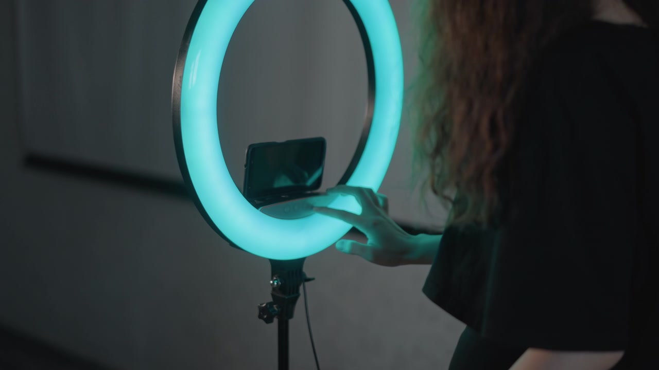 Rear three-quarter view shows person with long curly hair leaning forward, raising arm to adjust smartphone mount inside glowing ring light, with blue, cyan, magenta accent illumination and soft
