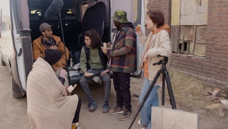 a movie production team taking a break while eating a sandwich and talking behind a van in the street