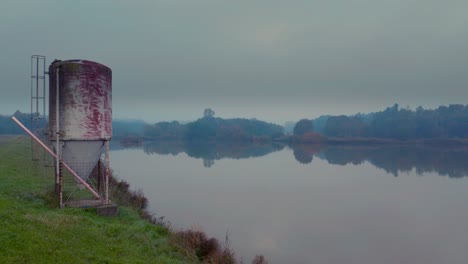 Aerial-view-reflective-foggy-lake-with-an-empty-metal-silo-on-a-grassy-hill