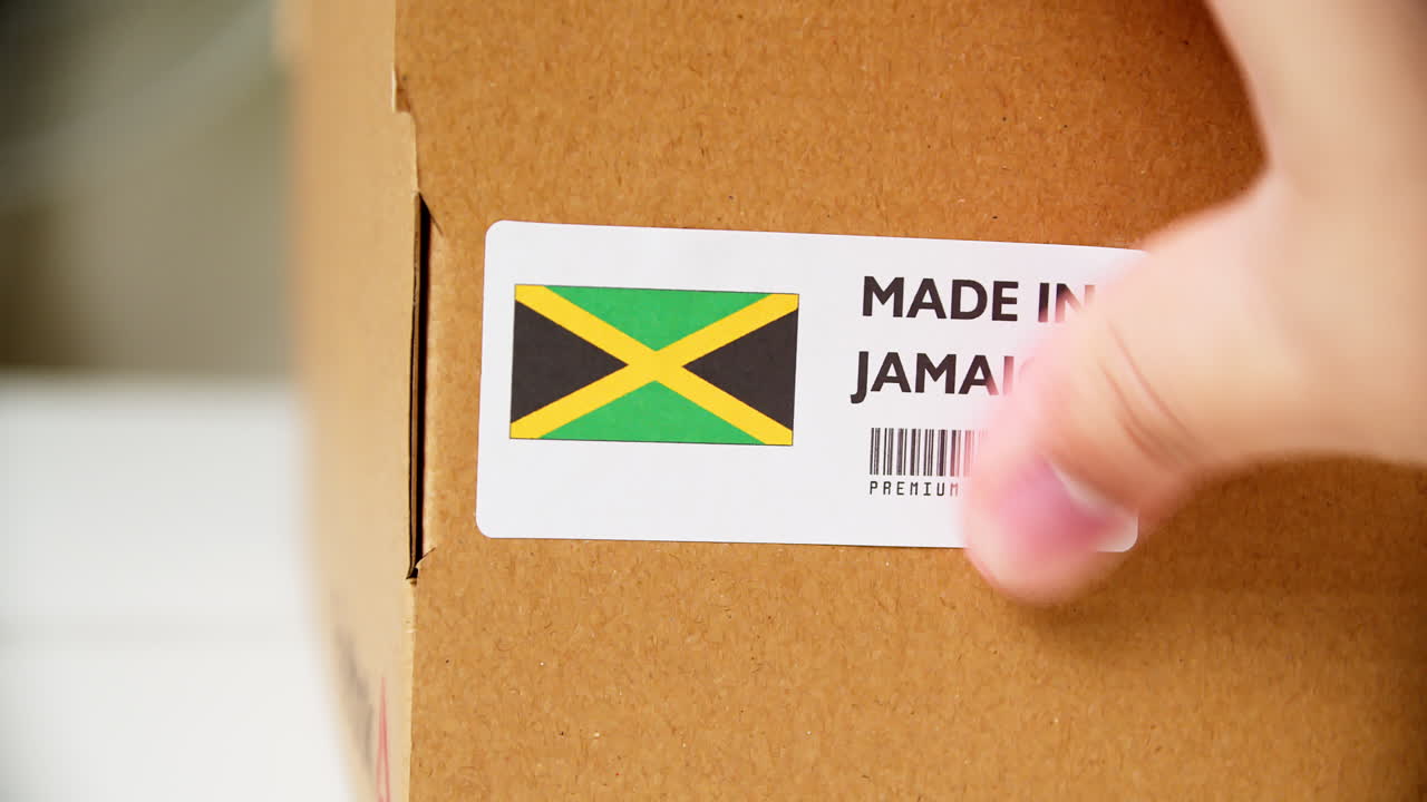 Hands applying MADE IN JAMAICA flag label on a shipping cardboard box with products. Close up shot with premium quality barcode