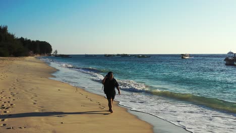 Woman-taking-photographs-with-smartphone,-walking-on-quiet-exotic-beach-alongside-sea-waves-on-a-bright-morning-sky-in-Malaysia