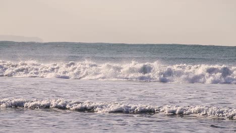 Die-Sanften,-Rollenden-Wellen-Am-Strand-Von-Carcavelos-Schaffen-Bei-Sonnenaufgang-Ein-Perfektes-Bild-Mit-Den-Leuchtenden-Farben-Des-Himmels-Und-Der-Kleinen,-Leuchtenden-Sonne,-Die-Einen-Blauen-Farbton-über-Das-Meer-Wirft