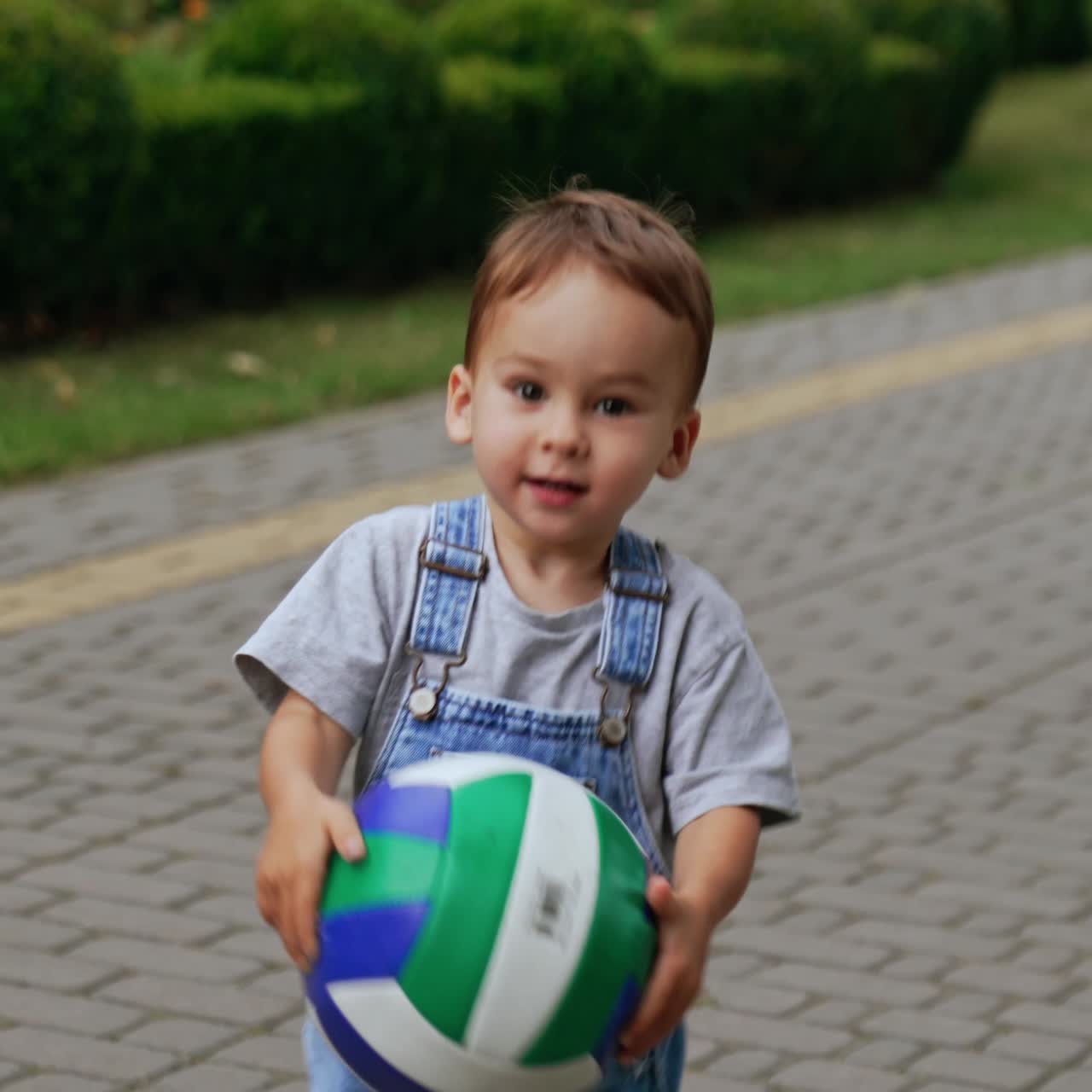 Caucasian toddler in jeans romper playing with a ball in the park. Happy child picks up a ball and runs with it approaching camera