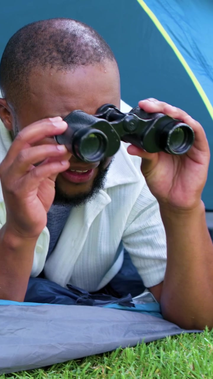 Vertical video: Smiling man in shirt reaching for black binoculars from companion at tent scanning