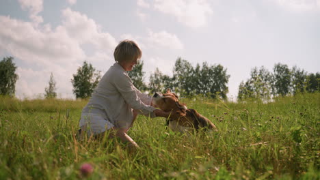 una mujer en cuclillas al aire libre frota el cuello de su perro mientras el perro agita las orejas felizmente en un campo cubierto de hierba rodeado de naturaleza y árboles en el fondo, capturando el vínculo lúdico y afectuoso entre la mascota y el dueño