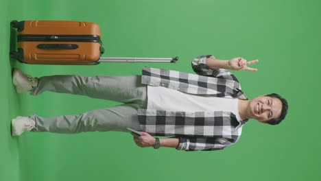 full body of asian male traveler with luggage and passport smiling and showing peace gesture while standing in the green screen background studio