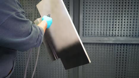 man applying black anti-corrosion powder coating to metal sheet using a spray gun in factory