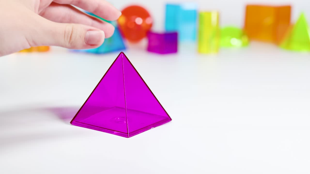 A hand interacts with vibrant geometric shapes on a white table, showcasing a purple pyramid among colorful prisms and spheres