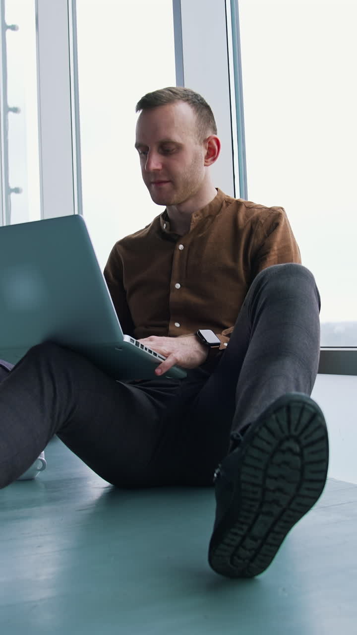 Young freelancer working on a laptop. Man in shirt and trousers sitting on a floor and using a gadget to work in office. Panoramic windows view. Vertical video