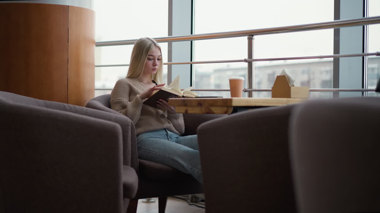 테이블에 앉아서 책을 읽고 두 손으로 고 있는 여자, 티슈 상자 의 나무 테이블 위에 커피 컵. 도시 풍경 전망으로 뒤에 큰 창문