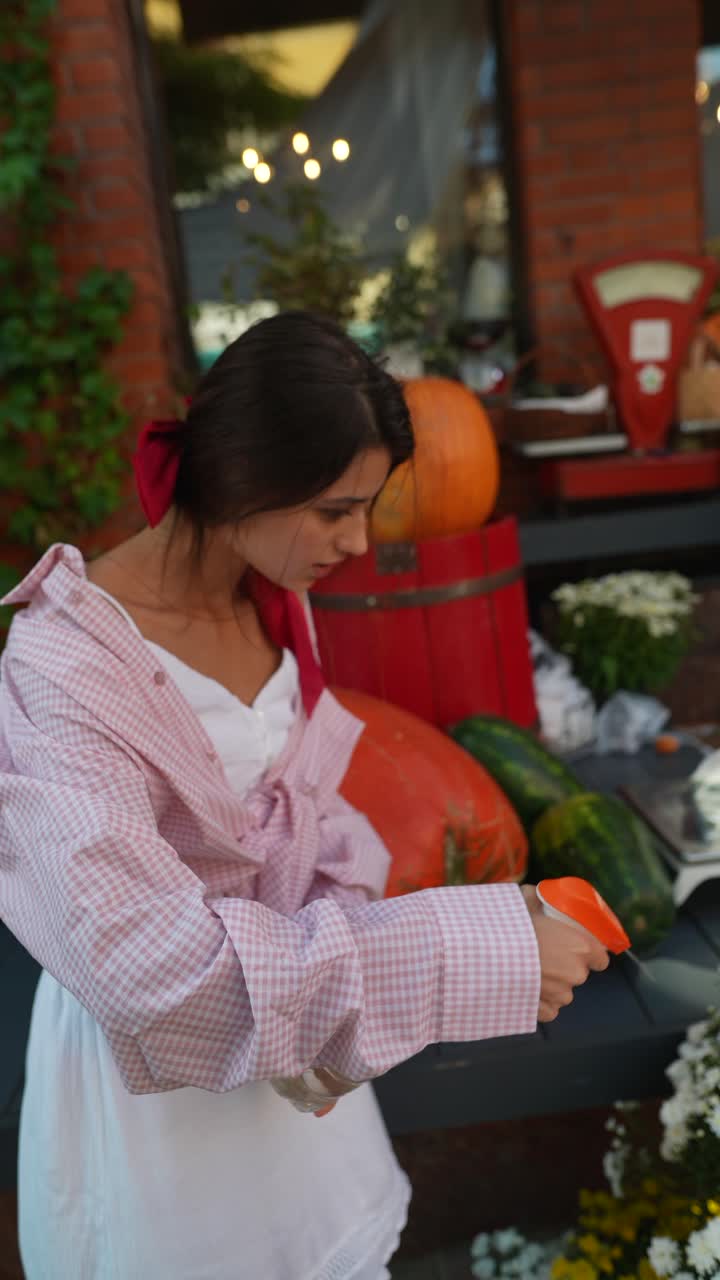 mujer con camisa a cuadros rosa y vestido blanco, al aire libre con calabazas y flores