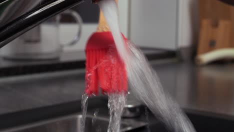 close up handheld shot of cleaning an oil brush under running water at the sink in the kitchen in slow motion