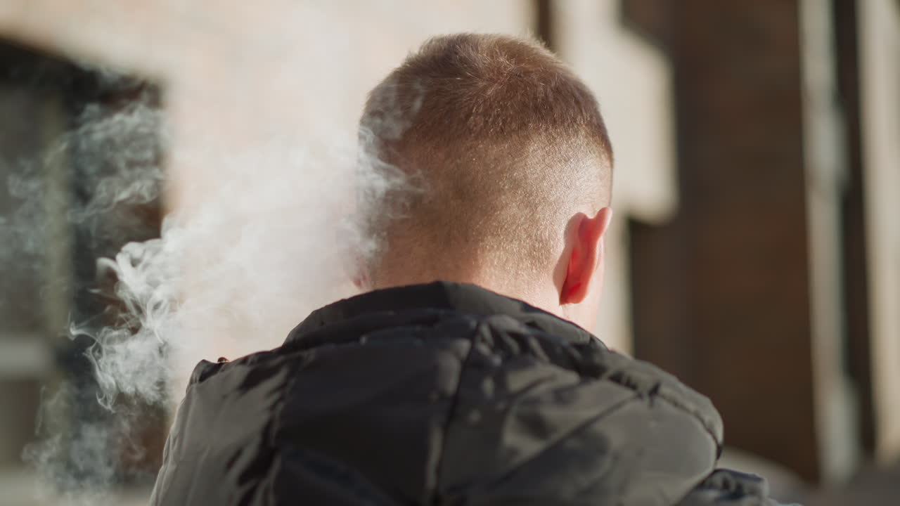 back view young man inhaling shisha outdoors on sunny day releasing thick smoke against blurred urban background with sunlight glow on jacket in vibrant warm late afternoon light soft haze