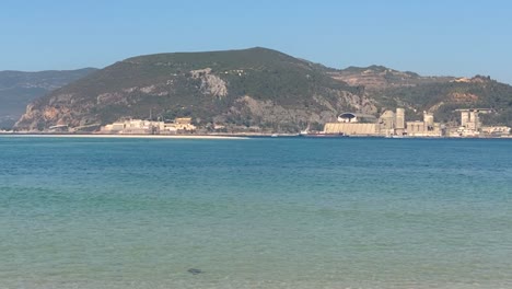 Wide-shot-of-coastal-city-on-top-of-cliffs-with-green-water-washing-sands-below-and-people-sailing-in-boats,-Portugal