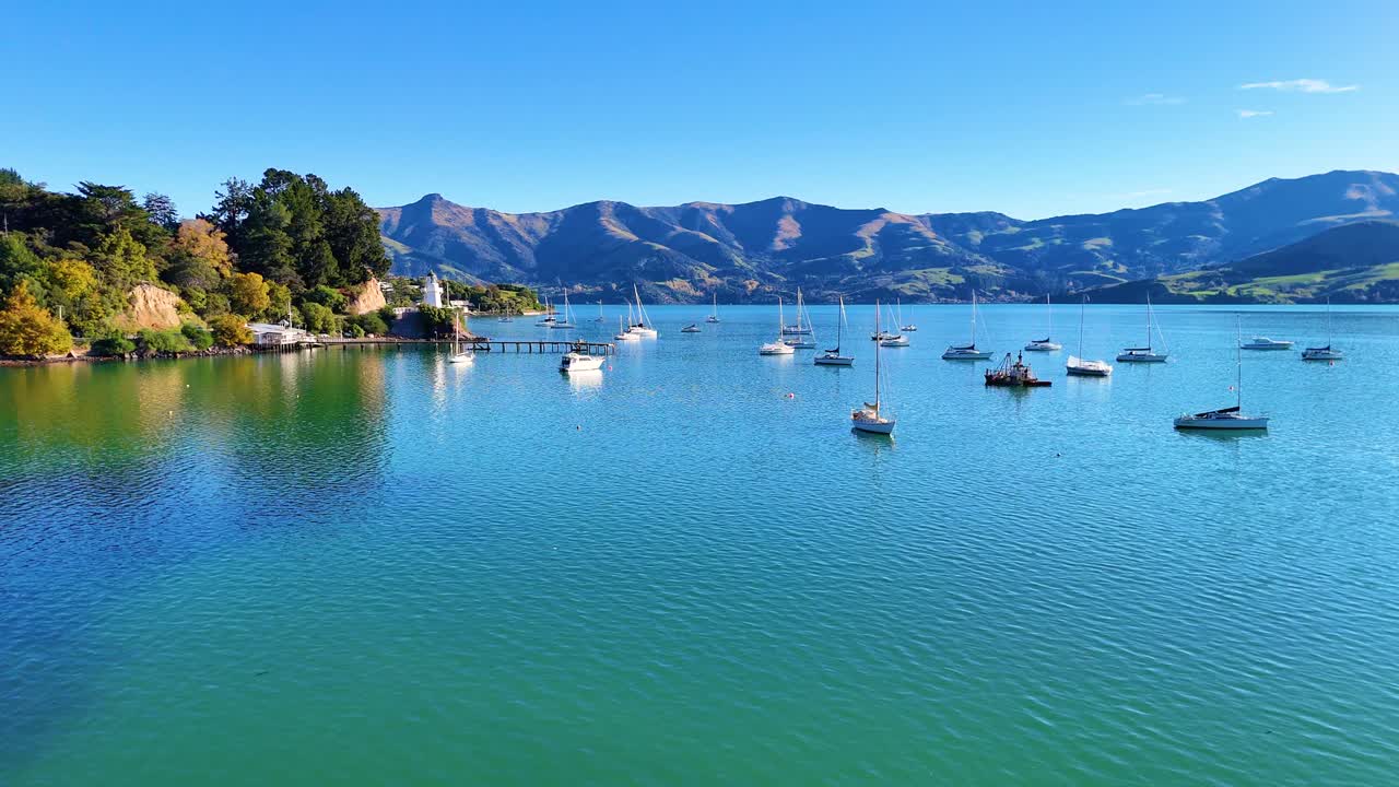 Calm harbor scene with anchored boats, clear skies, and distant hills in Akaroa, New Zealand. Bright daylight enhances tranquil atmosphere