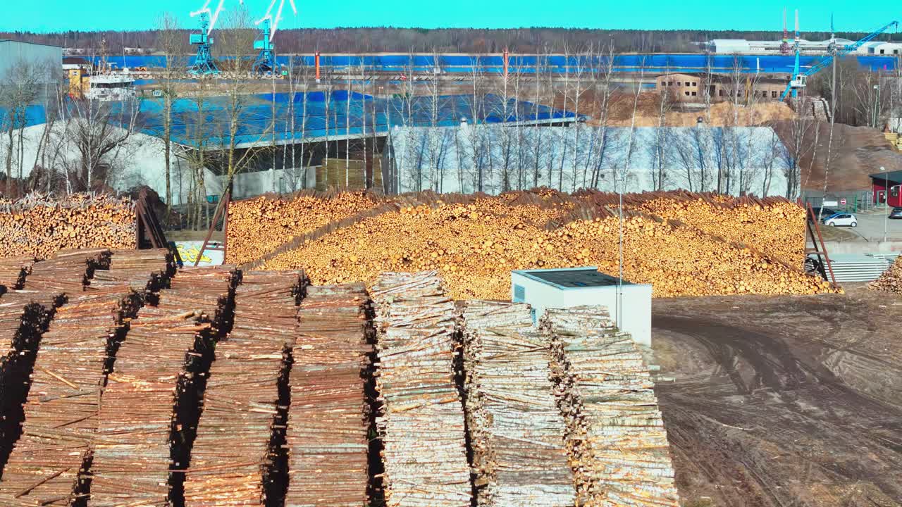 Latvian timber processing facility: vast stockpiles of logs in brown and tan hues under a hazy, overcast sky, viewed from a high-angle with a wide-angle lens, showcasing industrial scale