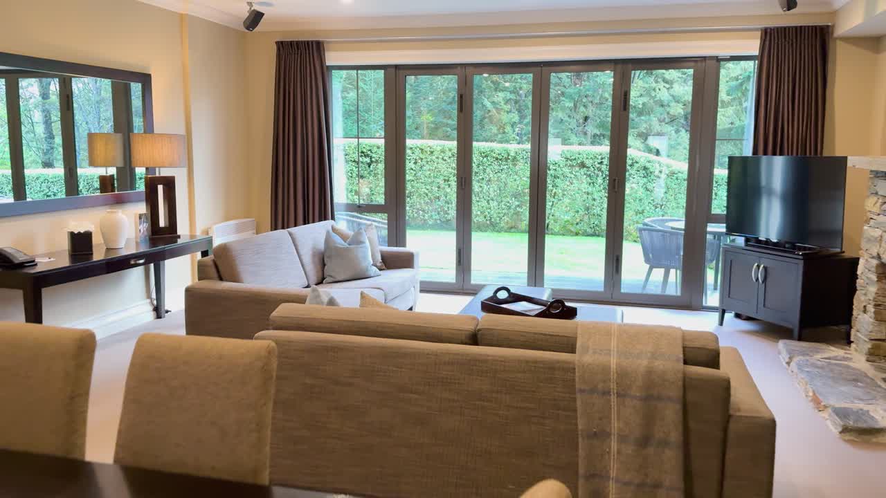 Camera smoothly pans through a bright, contemporary living and dining area with large windows, neutral furnishings, and lush garden scenery outside