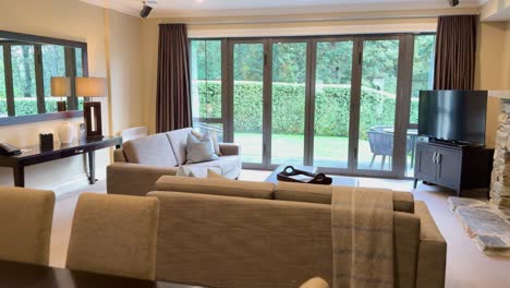 camera smoothly pans through a bright, contemporary living and dining area with large windows, neutral furnishings, and lush garden scenery outside