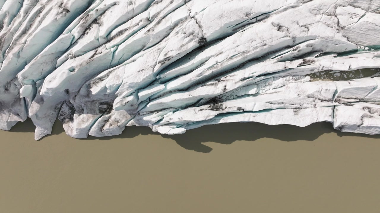 Detailed aerial view of the jagged front of Fjallsjökull Glacier meeting the calm, muddy waters of Fjallsarlon Glacier Lagoon in Iceland.