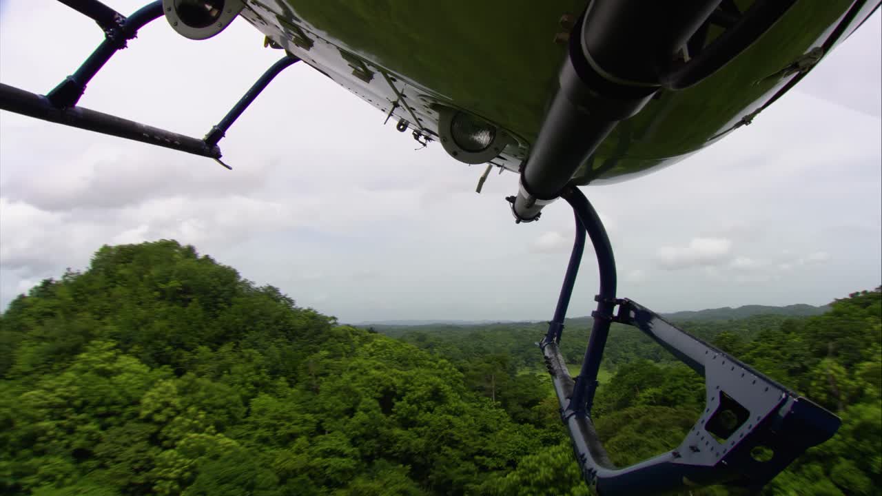 View from a helicopter in flight over the tropical jungle of El Petén, Guatemala. The aircraft’s fuselage and landing gear frame the lush forest landscape during a cinematic aerial recording mission.