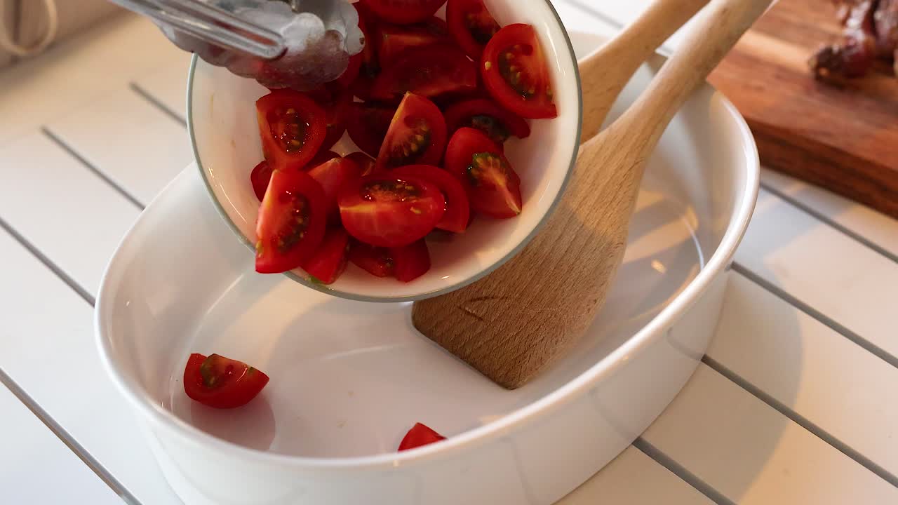 Cherry tomatoes are poured into a white bowl using tongs, with wooden utensils visible. Bright, natural lighting enhances the scene