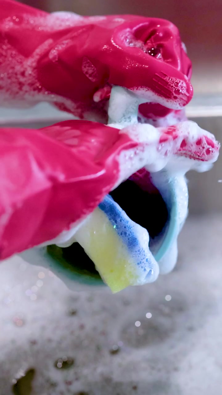 Close-up of hands in pink gloves cleaning a cup with a sponge in soapy water