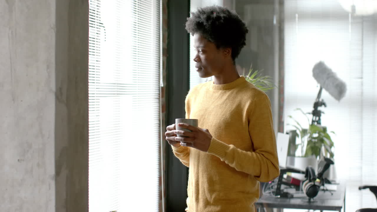 Thoughtful african american man drinking coffee and looking through window at home, slow motion