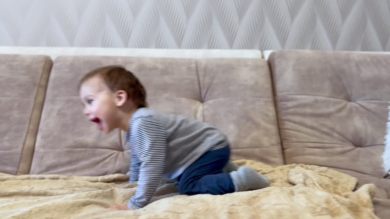 Beautiful active Caucasian toddler having fun jumping on the sofa. Kid waves his hands excitedly and smiles adorably.