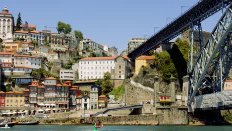 Central-Porto-Boats-passing-under-Luís-I-Bridge-with-Crowd-in-Background,-Portugal-4K-CINEMATIC-SUMMER-MEDITERRANEAN-CITY-RIVER-BANK