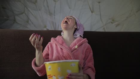 woman watching a late night movie at tv, eating popcorn. bathrobe, facial mask