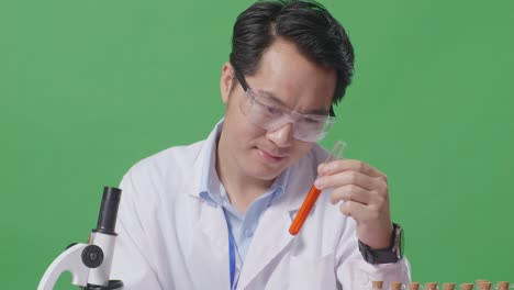 close up of asian man scientist looking at test tube then smiling and showing thumbs up gesture while working on the table with microscope in the green screen background laboratory