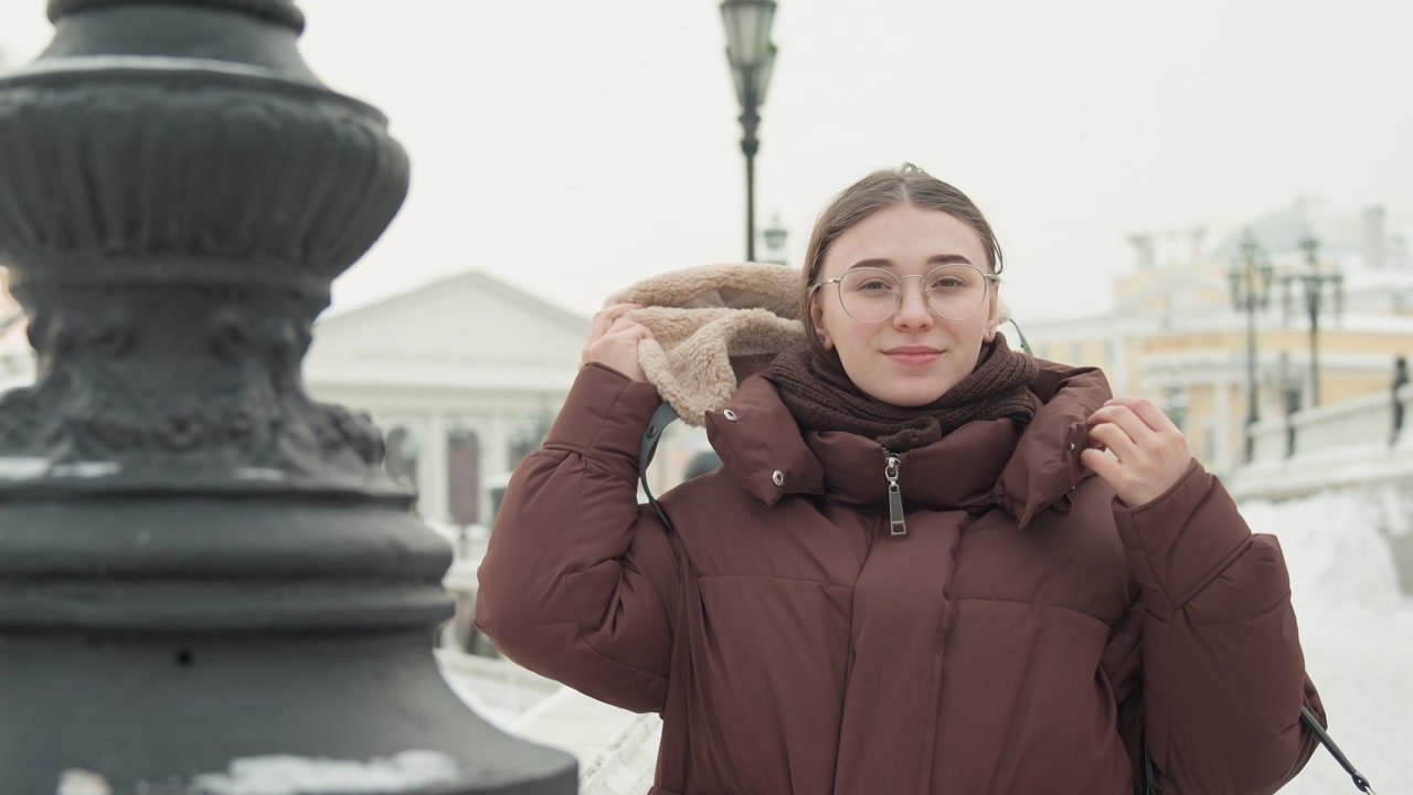 Giovane donna che indossa un piumino marrone con cappuccio bordato di pelliccia e occhiali, tiene un cappello di lana all'altezza del petto mentre alza le mani per sistemare gli strati invernali in un parco urbano innevato, con espressione calma e neutra.