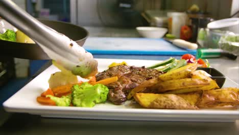 chef using metal tongs to place grilled vegetables on plate with steak and potatoes