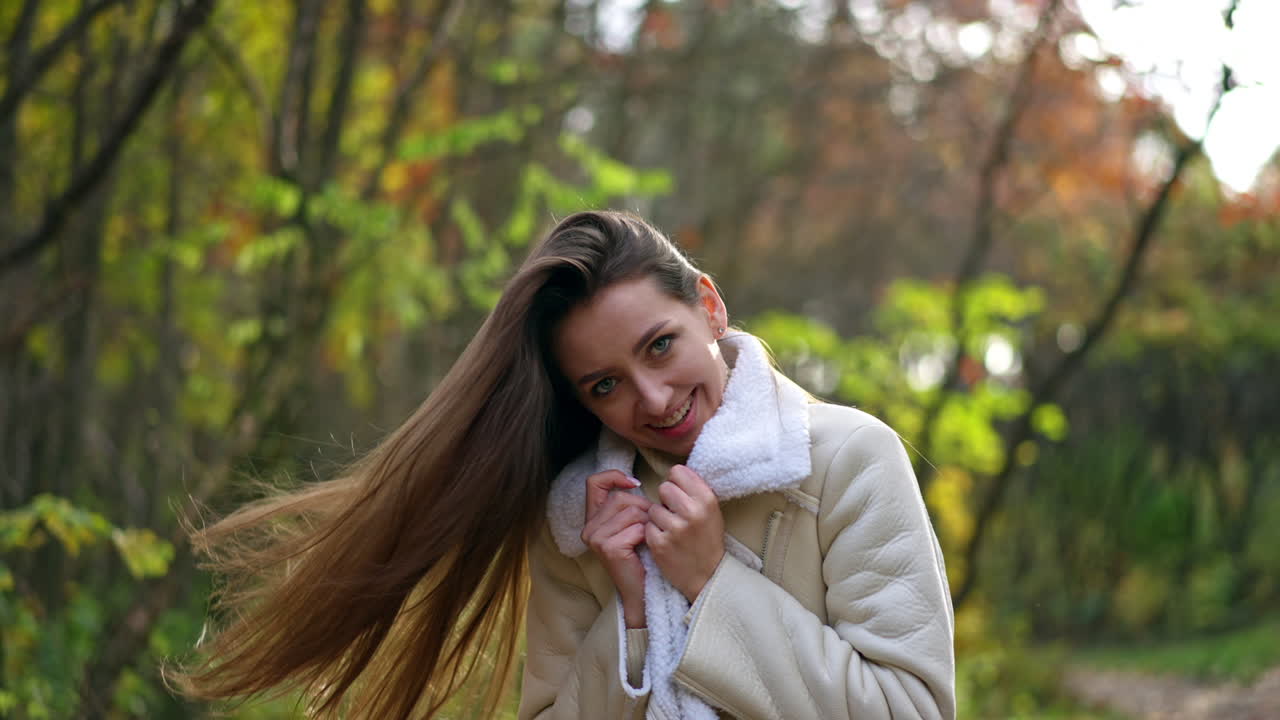 Brunette lady waves her long hair in front of camera. Positive smiling woman wearing warm clothes enjoying autumn weather.