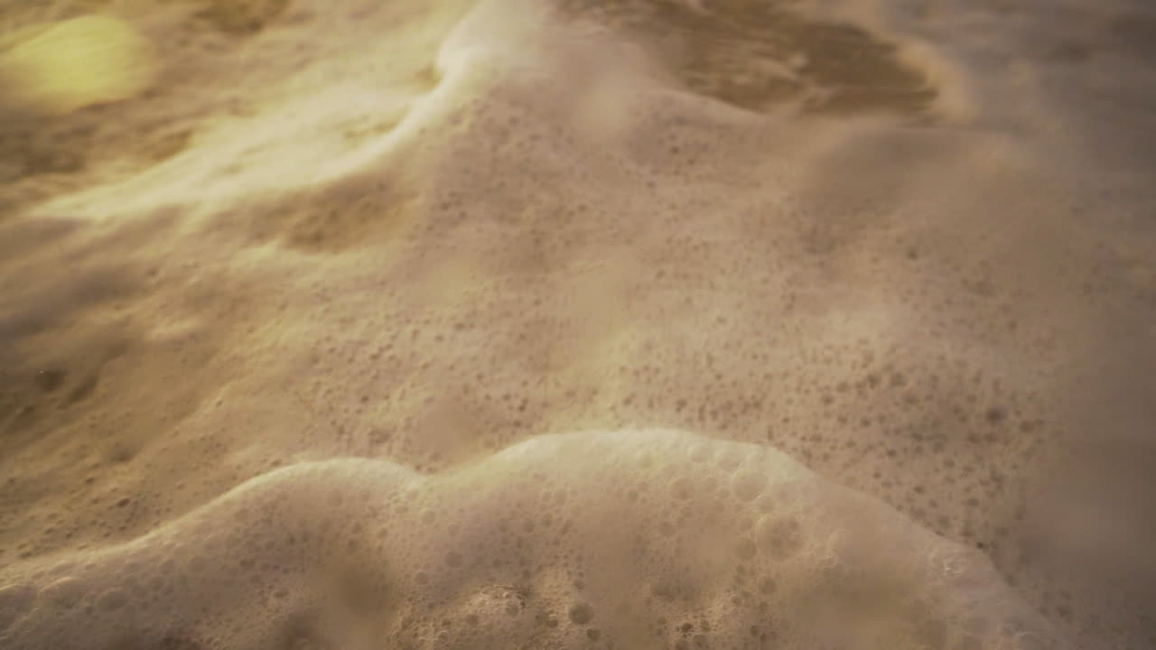 Close up shot of foam frothing waves on shore and then dives into the water and then rises, capturing a stunning view of the sunset over the ocean waves.