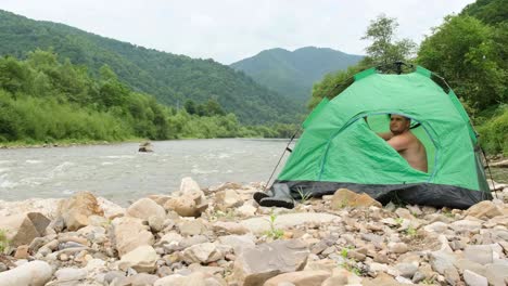 a young tourist opens a green tent on the bank of a mountain river and waves his hand a young man opens a green tent on the bank of a mountain river and waves his hand satisfied tourist