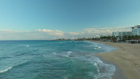 Aerial---panoramic-view-of-a-bustling-beachfront-in-Cyprus,-with-gentle-waves-lapping-the-sandy-shore