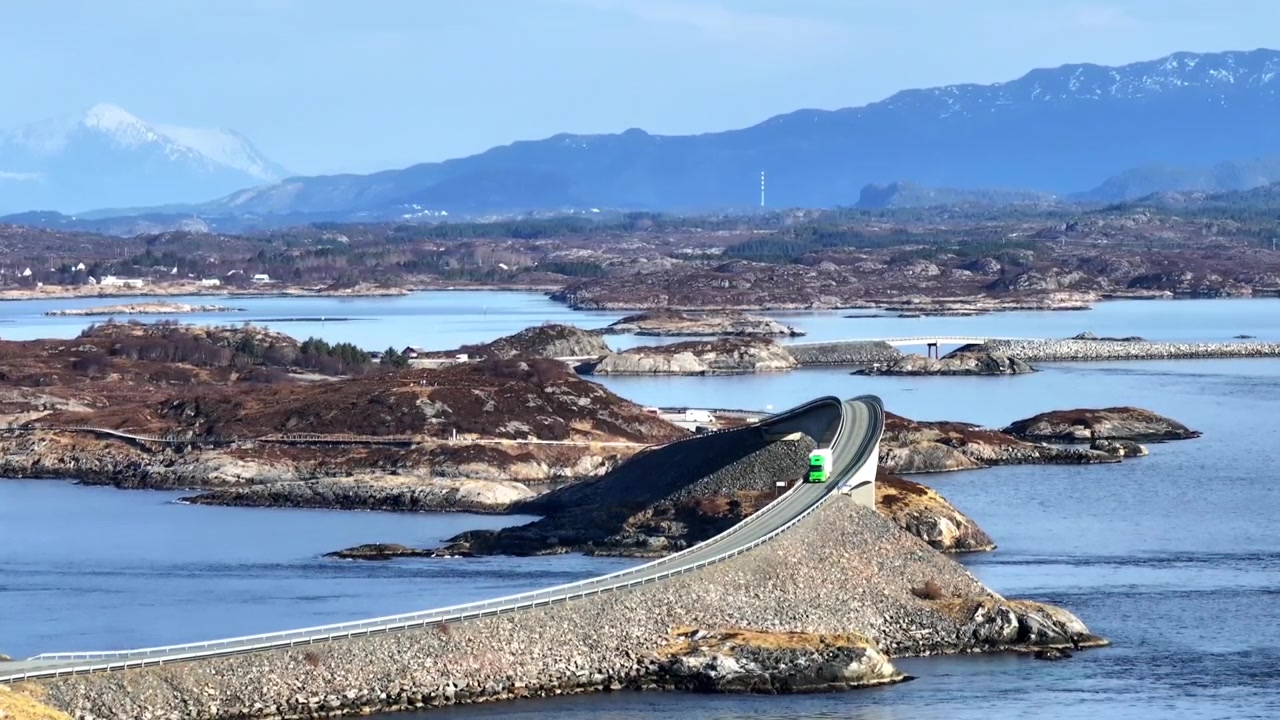 Vehicles cross a low coastal bridge between rocky islands with mountains in the distance.