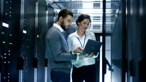 el especialista masculino muestra información en una computadora portátil al técnico de servidores femenino. están de pie en el centro de datos.